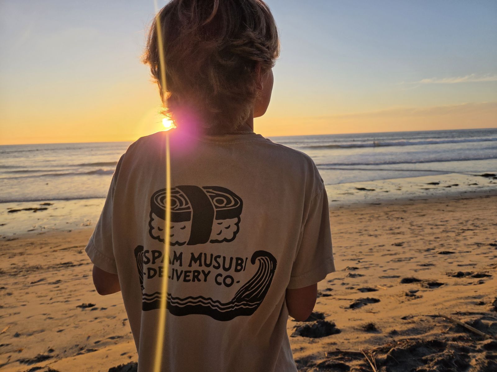 Young person facing a sunset on a beach, wearing a DTF printed "Spam Musubi Delivery Co." shirt. How Long Do DTF Prints Last?