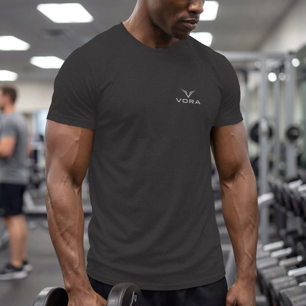 Man in a gym wears a dark Vora t-shirt, holding a dumbbell. The setting is modern and focused, with other gym-goers and equipment visible in the background.
