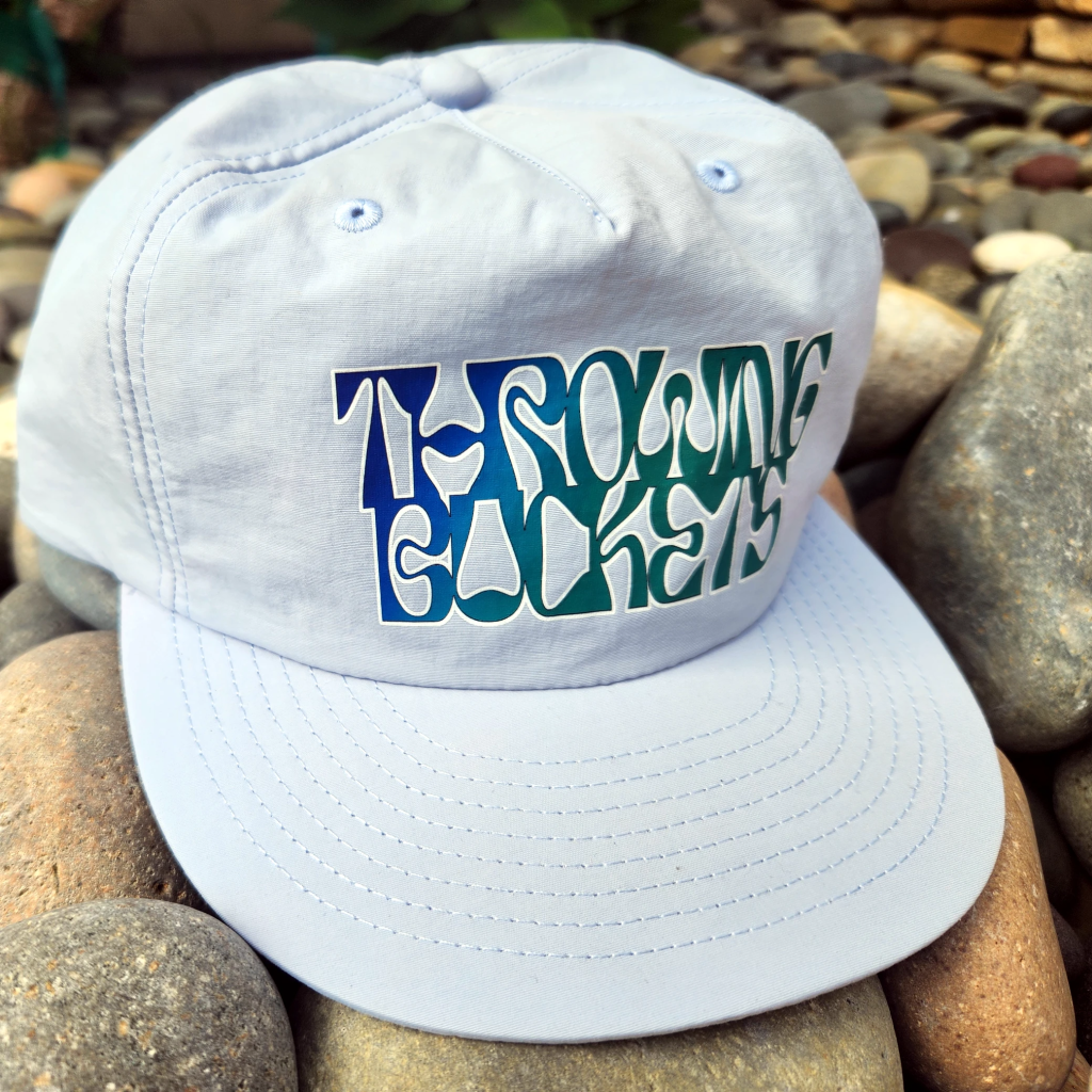 Light gray 1114 surf cap with “THROWING BUCKETS” in bold blue and green lettering, casual trendy style.