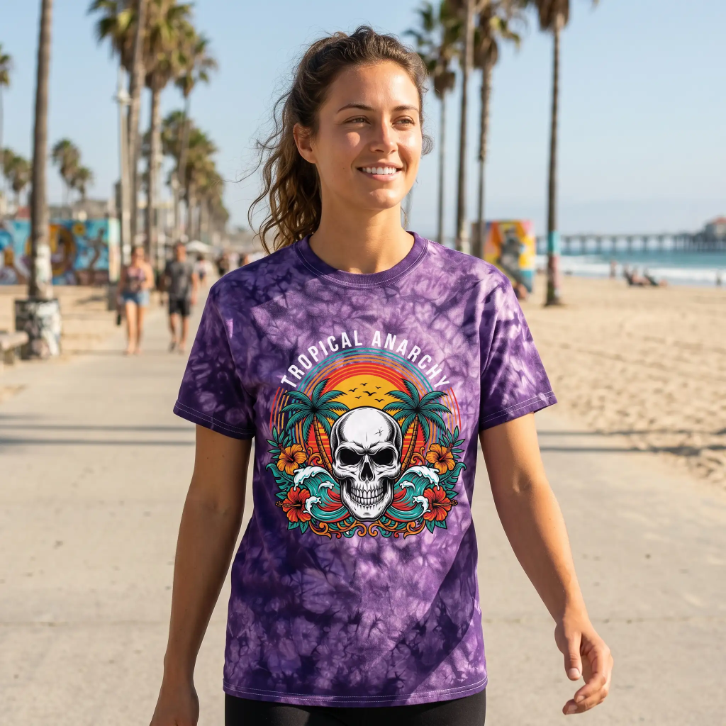 Smiling woman walking on a sunny beach promenade wearing a purple tie-dye shirt with a skull, palm trees, and "Tropical Anarchy" text.