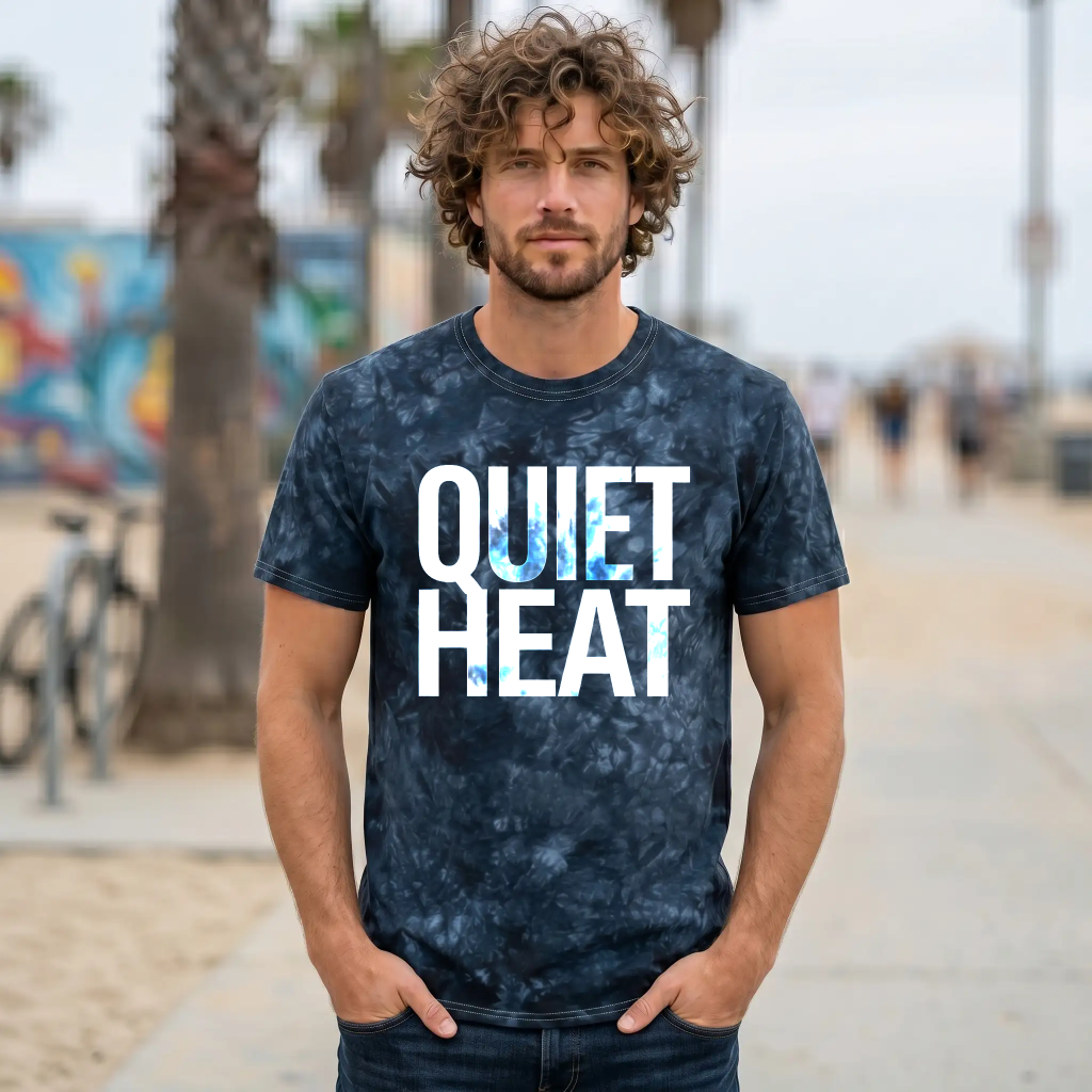 Man with curly hair wearing a navy dark tie-dye t-shirt (1390) standing confidently on a beach boardwalk with palm trees and graffiti in the background.